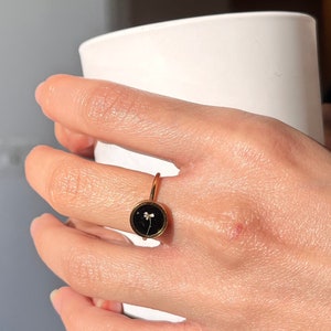May include: A gold ring with a black circular setting featuring a small white flower pressed inside.