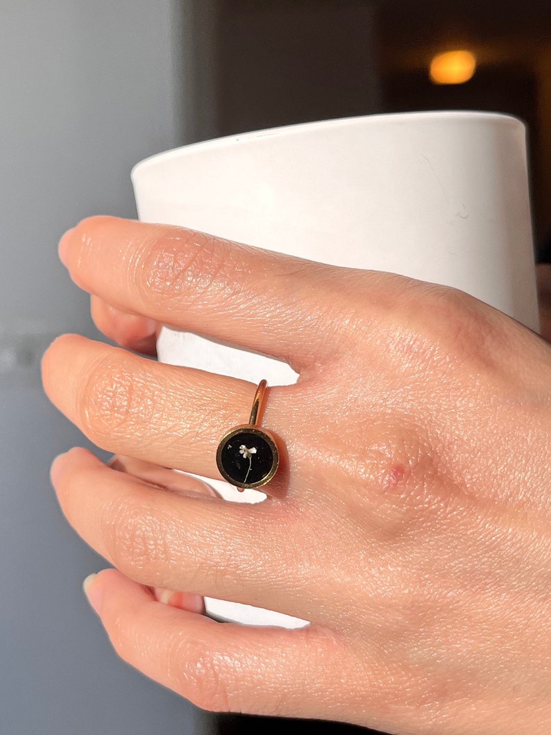 May include: A gold ring with a black circular setting featuring a small white flower pressed inside.