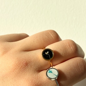 May include: Two gold rings with a round bezel setting. The top ring has a black background with a small white flower. The bottom ring has a white background with a blue flower.