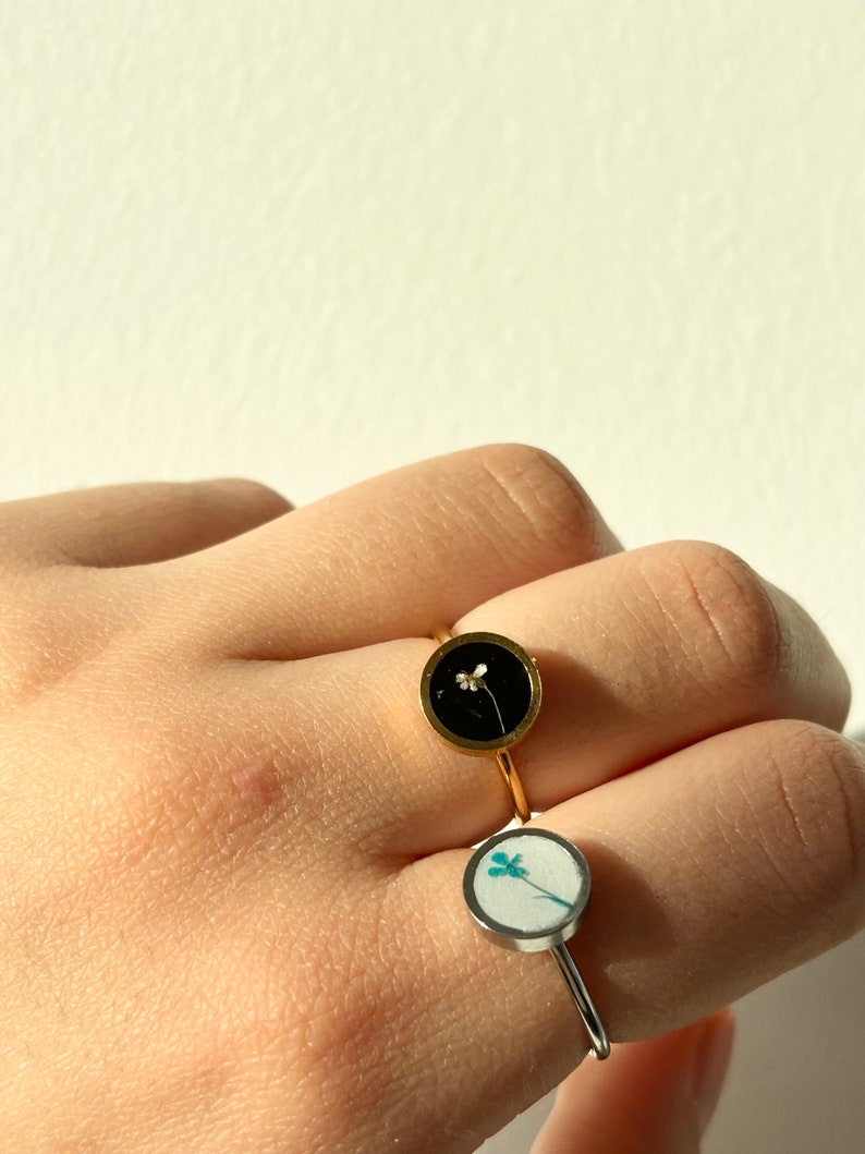 May include: Two gold rings with a round bezel setting. The top ring has a black background with a small white flower. The bottom ring has a white background with a blue flower.
