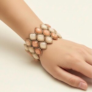 May include: A gold-coloured bracelet with a geometric design. The bracelet features multiple rows of square-shaped, peach and cream-coloured stones. The bracelet is worn on a wrist, showcasing the jewellery's design and colour.