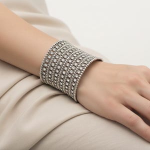 May include: Silver cuff bracelet featuring a repeating pattern of small, clear crystals. The wide bracelet is worn on a wrist. The design is visible against a neutral background.
