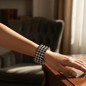 Peut inclure: Bracelet à plusieurs rangs avec des pierres noires et du métal argenté. Le bracelet est porté au poignet d'une personne, posé sur une surface en bois. Les pierres sont rondes et rapprochées.
