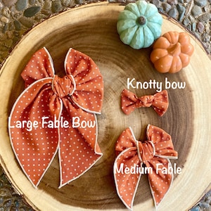 May include: Three orange and white fabric bows with a white trim. The largest bow is labeled "Large Fable Bow", the medium bow is labeled "Medium Fable", and the smallest bow is labeled "Knotty Bow".