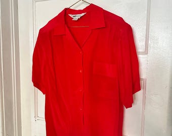 Pode incluir: Uma camisa vermelha de manga curta com colarinho clássico, um bolso no peito no lado esquerdo e uma fila de botões vermelhos a condizer na parte da frente. A peça de vestuário está pendurada num cabide de metal dourado.