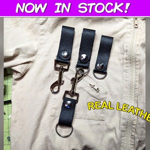 May include: Black leather keychains with silver-tone hardware, including snap closures and swivel clasps. The keychains are displayed on a tan fabric background. Text reads "NOW IN STOCK!" and "REAL LEATHER!"