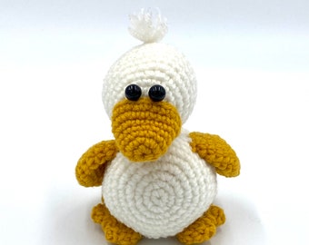 May include: A handmade crochet duckling toy. The duckling is primarily white with a yellow beak, feet, and wings. It has black safety eyes and a small white pom-pom on top of its head. The toy is designed with a rounded body and a friendly appearance.