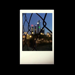 May include: A blurry image of a city skyline at night, seen through a chain link fence. The buildings are tall and slender, with lights shining from their windows. The fence is made of metal and is in focus, creating a stark contrast to the blurry background.