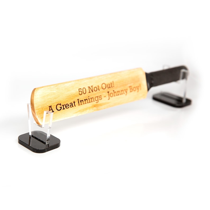 May include: A wooden cricket bat with the text "50 Not Out! A Great Innings - Johnny Boy!" is displayed on a clear stand with a black base. The bat has a black handle and a light brown blade.