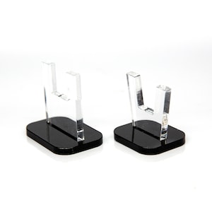 May include: Two clear acrylic display stands with a U-shaped top and a rectangular black base. The stands are designed to hold small items and are set against a white background.