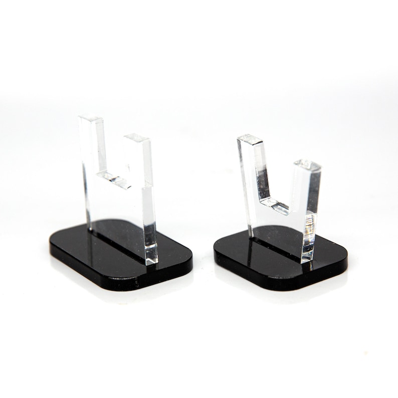 May include: Two clear acrylic display stands with a U-shaped top and a rectangular black base. The stands are designed to hold small items and are set against a white background.