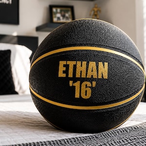 May include: Black basketball with gold stripes and the name "ETHAN '16" printed in gold. The ball is sitting on a bed with a white and gray patterned comforter. The background includes a window and a framed picture.