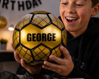 May include: A gold and black soccer ball with the name "GEORGE" printed in large black letters. The ball has a geometric pattern and is being held by a person.
