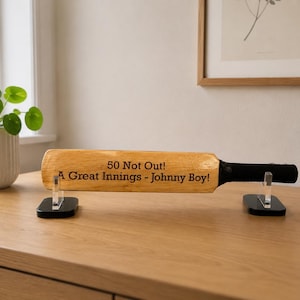 May include: A wooden cricket bat with the text "50 Not Out! A Great Innings - Johnny Boy!" is displayed on a wooden surface. The bat is supported by two clear acrylic stands with black bases. The handle is black.