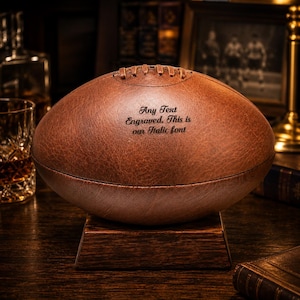 May include: A brown leather football with the text "Any Text Engraved. This is our Italic font" is displayed on a wooden stand. The football has visible stitching and is set against a dark, vintage-themed background.