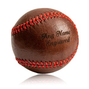 Puede incluir: Una pelota de béisbol de cuero marrón con costuras rojas. La pelota tiene el texto "Any Name Engraved" en una fuente cursiva. La pelota está sobre un fondo blanco con un ligero reflejo.
