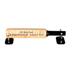 May include: A wooden cricket bat with the text "50 Not Out! A Great Innings - Johnny Boy!" is displayed on two black stands. The bat has a black grip and is made of light-colored wood.