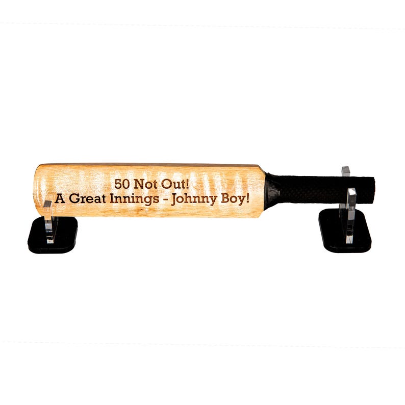 May include: A wooden cricket bat with the text "50 Not Out! A Great Innings - Johnny Boy!" is displayed on two black stands. The bat has a black grip and is made of light-colored wood.