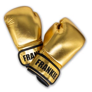 Puede incluir: Un par de guantes de boxeo dorados con correas negras para la muñeca. La palabra "FRANKIE" está impresa en negrita negra en cada correa. Los guantes están hechos de un material brillante y metálico.