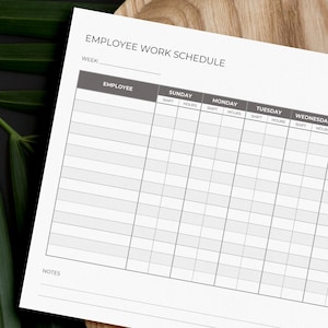 May include: A white employee work schedule with gray headings and a grid layout, featuring the text "Employee Work Schedule" and a logo. The schedule is on a wooden tray, with green leaves in the background.