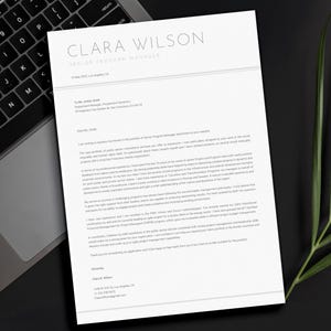 以下が含まれることがあります： 「CLARA WILSON SENIOR PROGRAM MANAGER」と題された白い履歴書が、ノートパソコンのキーボードと緑の葉の隣の黒い表面に表示されています。テキストは黒です。履歴書のタイトルは一番上にあります。