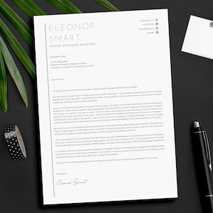 Può includere: Un curriculum vitae su carta bianca con il nome "Eleonor Smart" in alto. Una penna nera, un blocco per appunti, una clip e un rotolo di nastro adesivo a pois bianchi e neri sono su una superficie nera. Foglie verdi sullo sfondo.