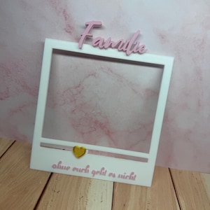 Op de afbeelding: Een witte fotolijst met het woord "Familie" in roze script bovenaan. De zin "ohne euch geht es nicht" staat in roze script onderaan, met een gouden hart. De lijst staat op een houten oppervlak.