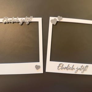 Op de afbeelding: Twee witte fotolijsten met grijze hartaccenten en de Duitse zin "Endlich jetzt!" in zilveren letters. Eén lijst heeft de tekst erboven, de andere eronder. De lijsten zijn vierkant met een rechthoekige opening.