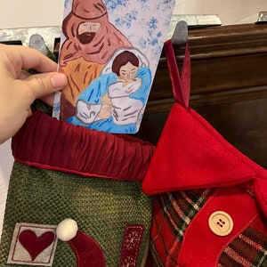 May include: A green and red Christmas stocking with a Santa Claus design and a red heart. A card with a watercolor painting of Mary, Joseph, and baby Jesus is tucked into the stocking.
