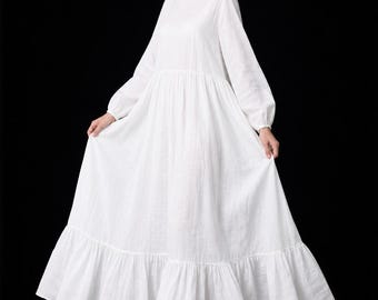 Könnte beinhalten: Ein langärmeliges weißes Maxikleid mit Rundhalsausschnitt, geraffter Taille und einem voluminösen Rock mit breitem Rüschensaum. Das Kleidungsstück besteht aus einem leichten, strukturierten Stoff, der in einer lockeren Silhouette bis zum Boden fällt.