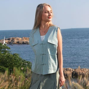 May include: A woman wearing a light blue sleeveless linen vest with a mandarin collar and a matching light green linen skirt. She is standing on a grassy cliff overlooking the ocean.