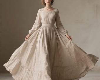 May include: A floor-length beige dress with long sleeves, a round neckline, and a fitted waist. The skirt features a full, flowing silhouette with a wide gathered tier at the bottom hem, crafted from a textured fabric that drapes to the floor.
