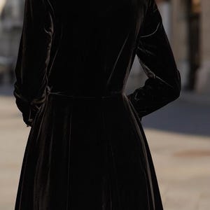 Puede incluir: Vestido largo de terciopelo negro con mangas largas y corpi&ntilde;o entallado. El vestido tiene una falda acampanada y un cuello alto. La modelo lleva pendientes peque&ntilde;os y redondos. El fondo est&aacute; borroso.