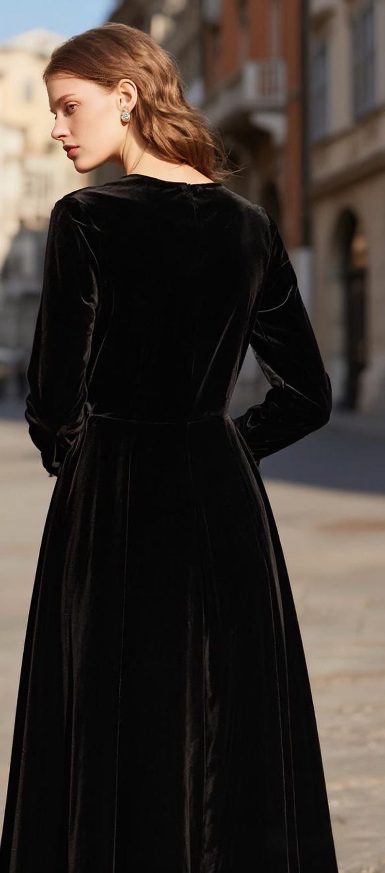 Puede incluir: Vestido largo de terciopelo negro con mangas largas y corpi&ntilde;o entallado. El vestido tiene una falda acampanada y un cuello alto. La modelo lleva pendientes peque&ntilde;os y redondos. El fondo est&aacute; borroso.