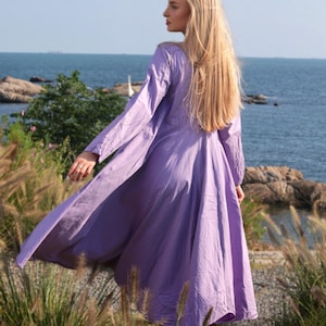 Pode incluir: Uma mulher com um vestido lavanda fluido est&aacute; em p&eacute; em um penhasco gramado com vista para o oceano. O vestido tem mangas compridas e uma saia fluida. O cabelo da mulher &eacute; longo e loiro.