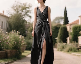 Puede incluir: Un vestido envolvente sin mangas, gris oscuro, con cuello en V y cinturón en la cintura. El vestido tiene una abertura alta en el lado izquierdo. La modelo camina por un camino de piedra, con vegetación y edificios al fondo.