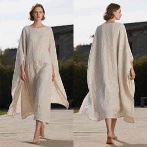 May include: A long, flowing, cream-colored linen dress with a round neckline and wide sleeves. The dress has a loose, comfortable fit and falls to the ankles. The model is barefoot and walking on a stone path.
