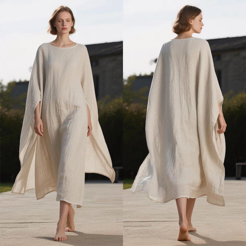 May include: A long, flowing, cream-colored linen dress with a round neckline and wide sleeves. The dress has a loose, comfortable fit and falls to the ankles. The model is barefoot and walking on a stone path.