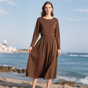 May include: A solid brown midi dress featuring long sleeves, a round neckline, and an elasticized waistline that transitions into a pleated, flowing skirt that falls below the knees. The garment is constructed from a smooth, plain fabric.
