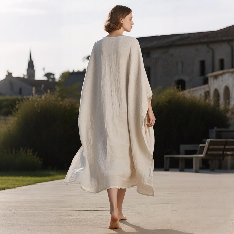 May include: A cream-colored, loose-fitting linen dress with a wide, flowing silhouette. The dress has a simple neckline and falls to the ankles. The model is walking on a concrete path in an outdoor setting.