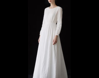 Puede incluir: Un vestido blanco largo hasta el suelo con mangas largas, cuello redondo y una cintura entallada que da paso a una falda amplia y fluida. La prenda presenta un tejido blanco liso y sencillo con una textura sutil y puños fruncidos en las muñecas.