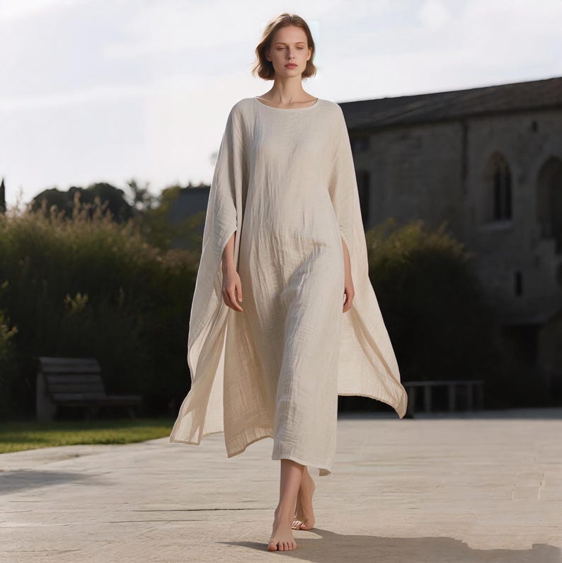 May include: A long, flowing, off-white linen dress with a round neckline and cape-like sleeves. The dress is ankle-length and has a relaxed fit. The model is walking on a stone path in front of a building.