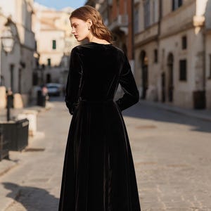 Puede incluir: Un vestido largo de terciopelo negro con mangas largas y una falda fluida. El vestido tiene un corpi&ntilde;o ajustado y un escote en V. La modelo est&aacute; de pie en una calle adoquinada frente a edificios.
