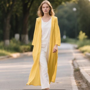 Könnte beinhalten: Ein langer, leuchtend gelber Kapuzenmantel, getragen über einem schlichten weißen ärmellosen Jumpsuit. Der Mantel hat lange Ärmel und ein offenes Vorderteil, während der Jumpsuit einen Rundhalsausschnitt und eine gerade geschnittene Hose besitzt.