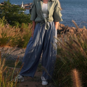 May include: A woman wearing a light green shirt tied at the waist and wide-legged blue pants. She is standing in a grassy area with a body of water in the background.