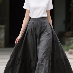 Pode incluir: Uma t-shirt branca de manga curta com gola redonda combinada com uma saia comprida maxi cinzento-escura. A saia apresenta um design de cintura subida com uma bainha franzida e em camadas que cai at&eacute; ao ch&atilde;o, criando uma silhueta volumosa.