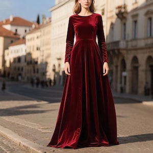 May include: A long, burgundy velvet dress with long sleeves. The dress has a round neckline and a flowing skirt. The dress is worn in an outdoor setting with a blurred background of buildings.