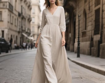May include: A floor-length, light beige maxi dress with a V-neckline, three-quarter length sleeves, and a gathered waistline. The garment features a textured fabric and a flowing, full-length skirt that reaches the ground, creating a simple silhouette.