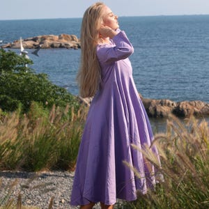 Pode incluir: Uma mulher com um vestido maxi lavanda est&aacute; em um campo de grama alta com o oceano ao fundo. Ela est&aacute; olhando para o lado e seu cabelo est&aacute; sendo levado pelo vento.