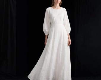 Puede incluir: Un vestido blanco largo hasta el suelo con mangas largas y voluminosas, escote redondo y un corpiño ajustado que se transforma en una falda amplia y fluida. La tela presenta un tejido fino y texturizado en toda la prenda.